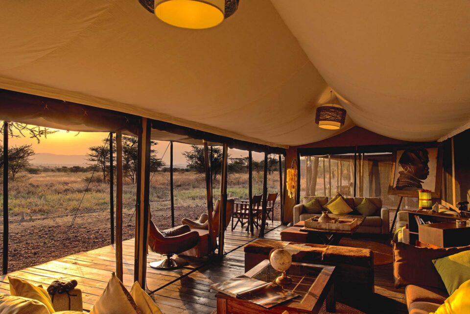 Inside a luxury tented safari camp at dusk, with comfortable lounge seating, warm ambient lighting, and unobstructed views of the African plains.